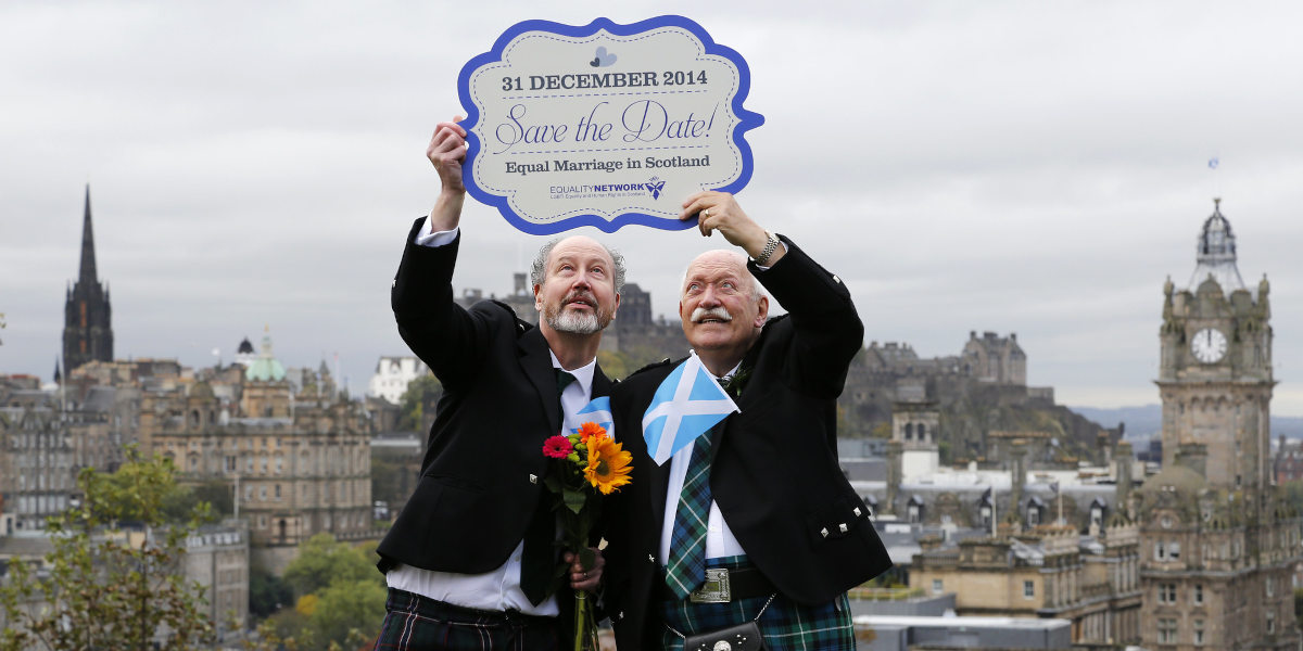 Two men stand in suits holding up a save the date message celebrating same-sex marriage laws