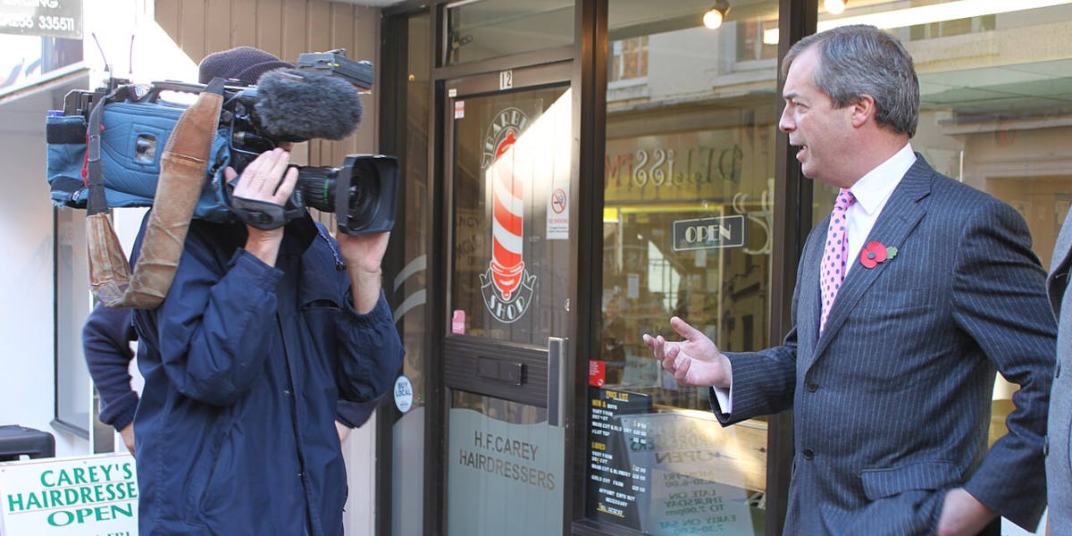 Reform UK (then UKIP) leader Nigel Farage (right) talking into a camera held by a cameraman (left) outside a shop