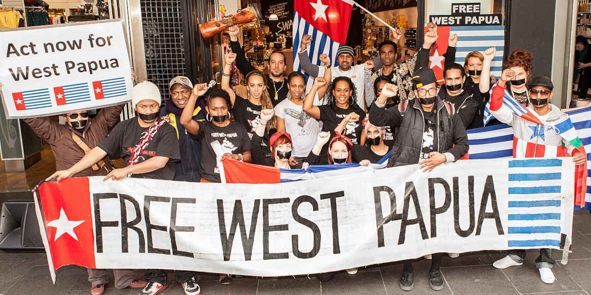 A group of people, some with raised fists or with tape over their mouths, standing behind a banner reading 'Free West Papua' and holding West Papuan flags