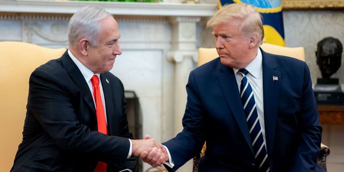 Two white men in suits shake hands while sitting on yellow chairs