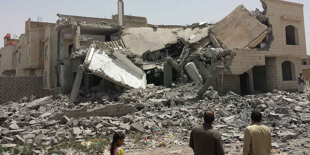 Three people looking on at a house that has been reduced to rubble in Sanaa, Yemen