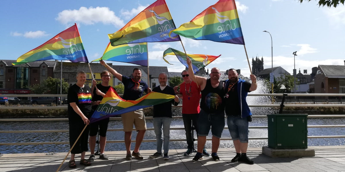 A group of people stand holding rainbow flags in the sun at Newry Pride