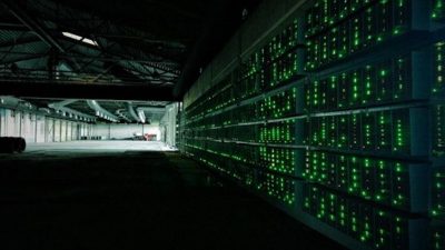 A photo showing bitcoing mining, showing a large, dark room full of computers displaying green lights