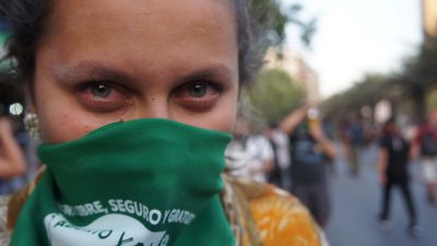 A person looks directly into the camera, a green scarf covering their nose and mouth