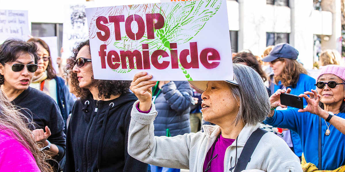 A woman with grey hair in a purple tee and grey jacket holds up a sign reading "STOP femicide" at a protest