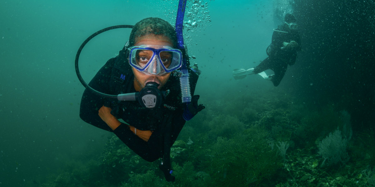A scuba diver underwater faces the camera, whilst another appears in the background