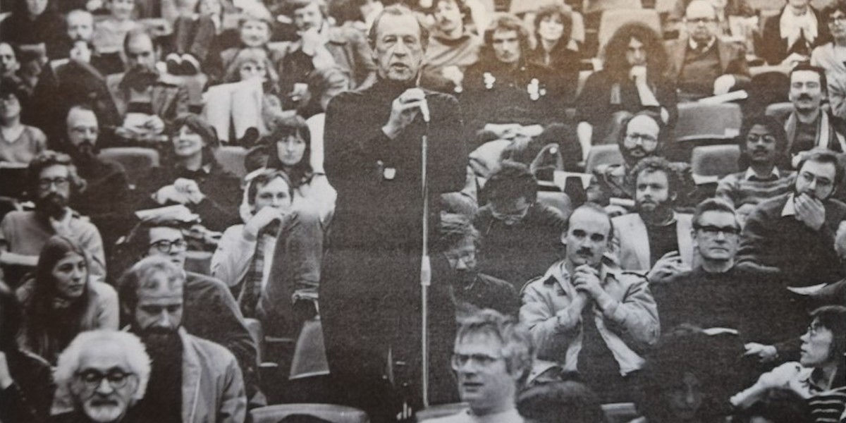 A man, Raymond Williams, ins standing up and speaking into a microphone surrounded by people seated all around him in a busy lecture hall