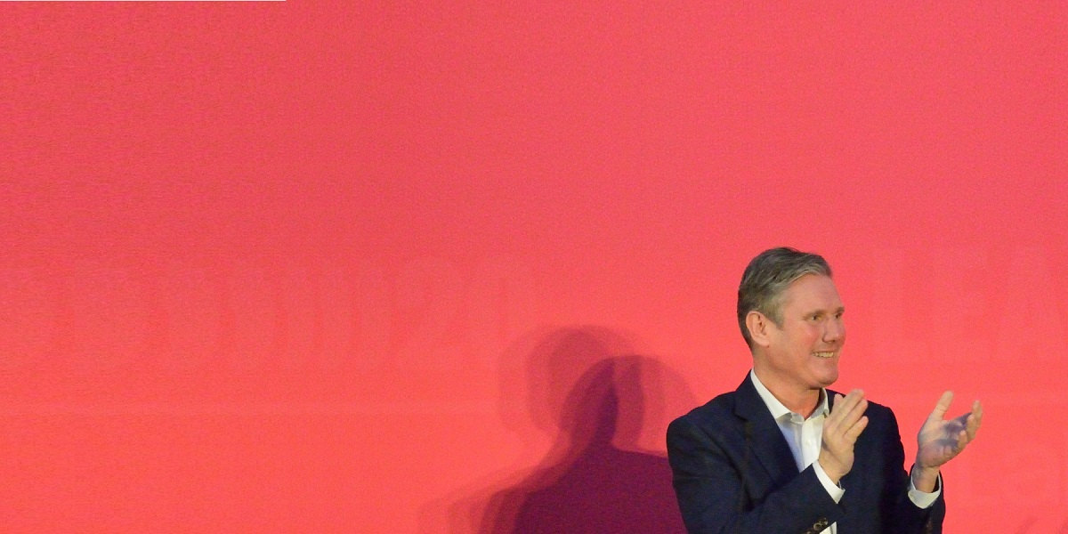 Labour leader Kier Starmer stands clapping at Party Conference against a red background
