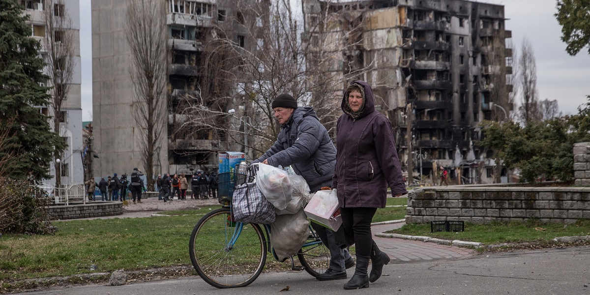 A woman and a man pushing a bicycle laden with bags in front of bombed blocks of flats