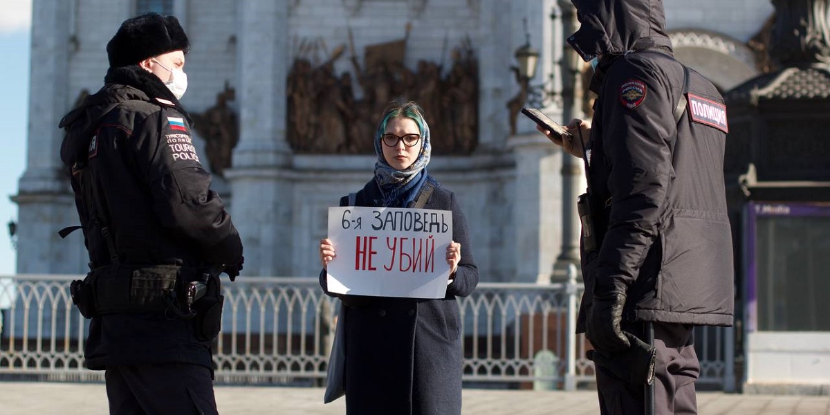 A woman holding a sign in Russian reading 'Sixth commandment – don't kill' flanked by two policemen