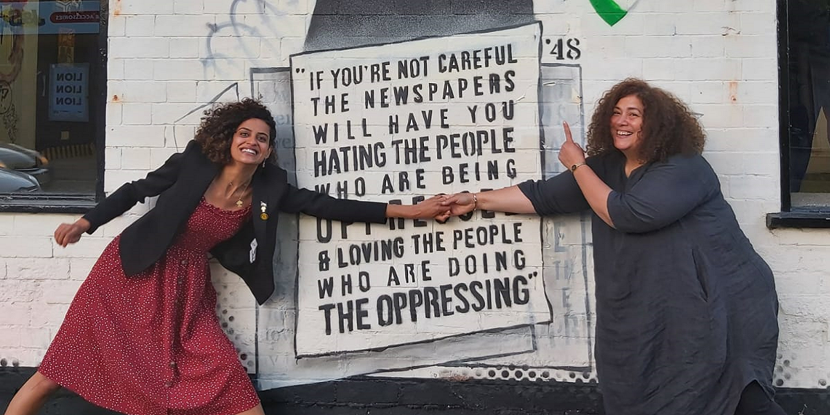 Shahd and her friend Shams, both smiling, with curly hair and wearing long dresses, hold hands across a mural of Malcom X made by Birmingham-based artist Mohammed Ali during the Israeli attack on Gaza in May 2021
