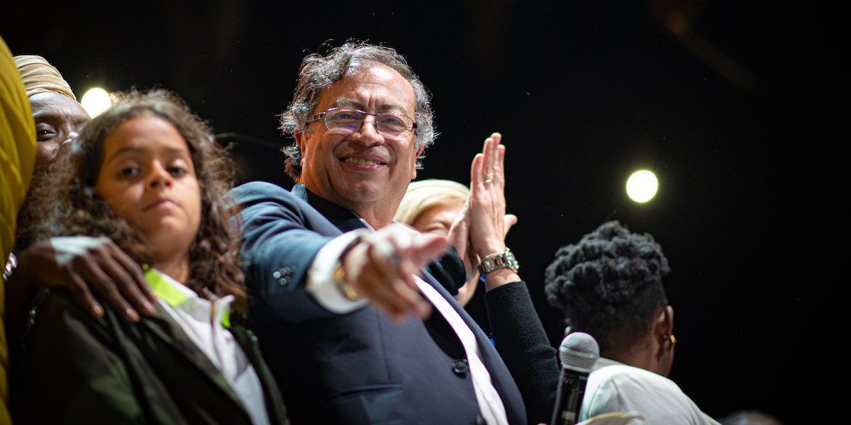 A man in a suit points at the camera, smiling, from a crowded stage