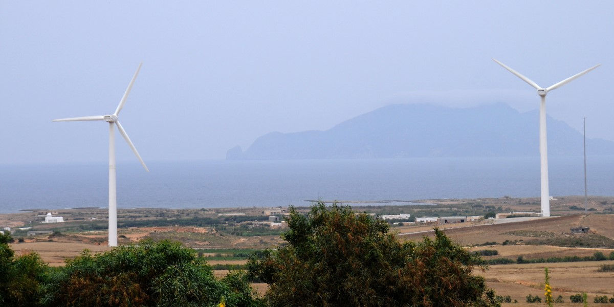 Two wind turbines in an arid landscape in the foreground, leading out to the sea and a distant mountainous island or peninsula