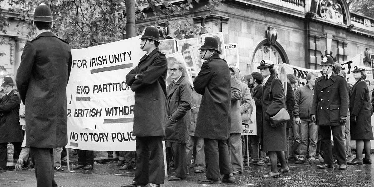 A protest march. Policemen in helmets and coats stand around a group of protesters. A banner reads 'for Irish unity, end partition, for British withdrawal, no to Tory policies'