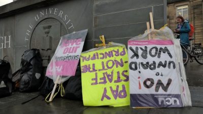 Upside placards supporting striking university and college workers are covered with clear plastic and lean against a grey wall during a rainy protest