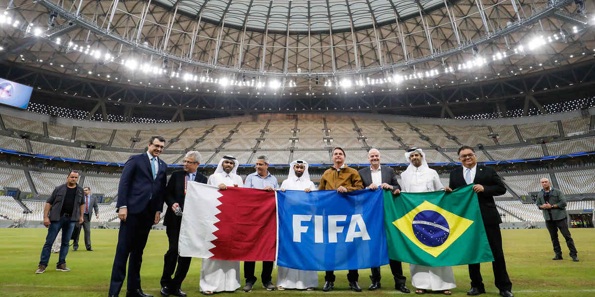 A row of men in western and Qatari dress stand on the grass in an empty stadium holding the flags of Qatar, Fifa and Brazil