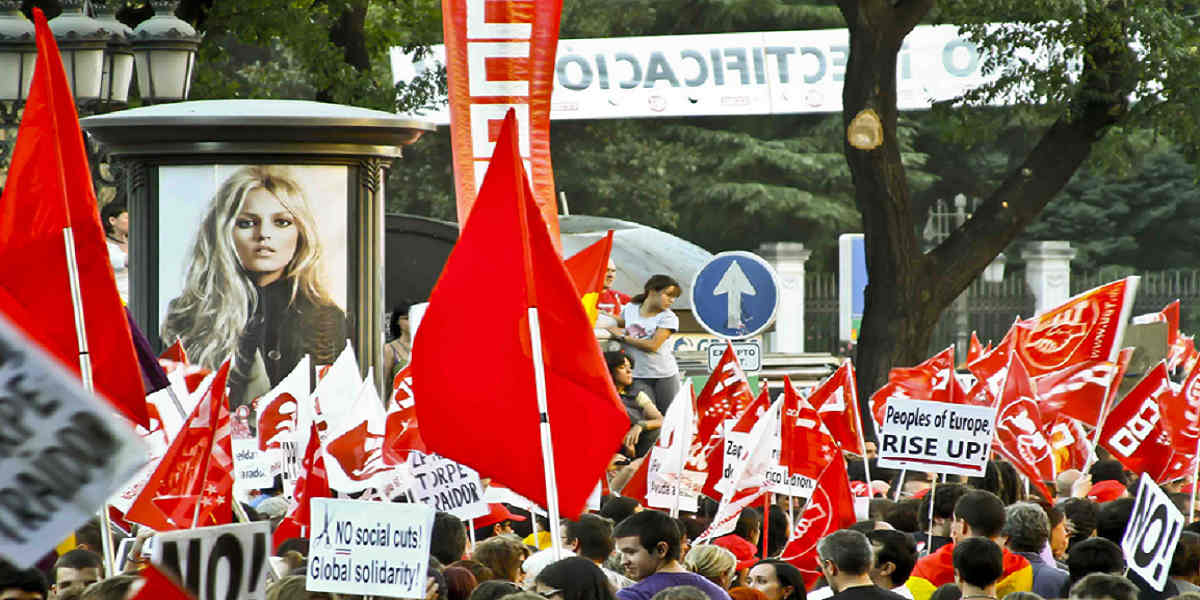 A protest march with banners and red flags. In the background an advertising column features a poster of a blonde model which seems to be staring back at the camera