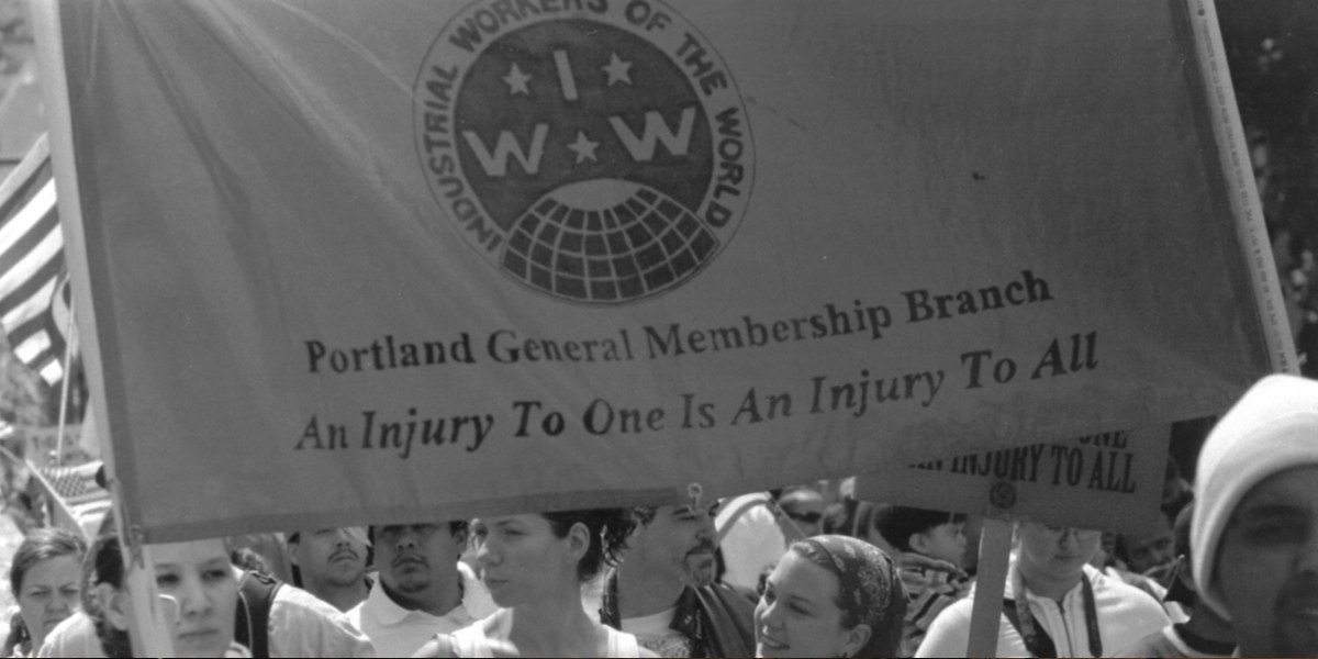 A protest march. A large banner is held above head height. It has the IWW logo and the words 'Portland General Membership Branch', 'An Injury to one is an injury to all'