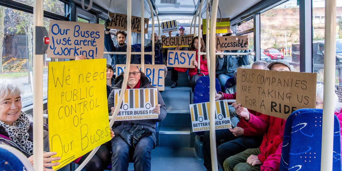 A group of people are sitting on the bus holding signs. The signs have handwritten slogans on them in support of public control of buses.