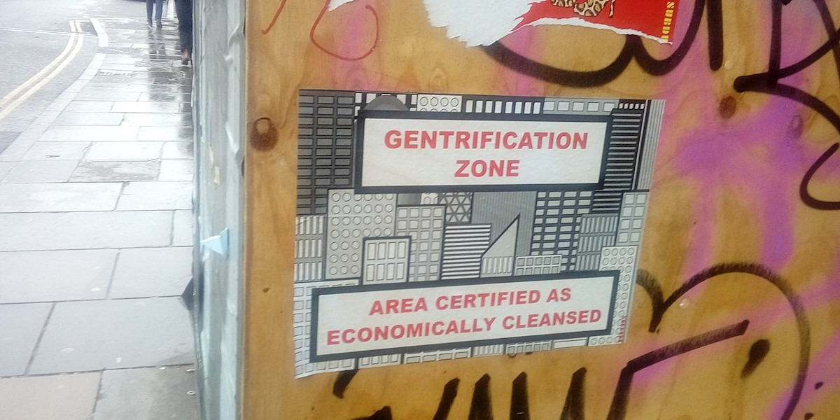On a city street building works hoarding, a poster reads: "Gentrification Zone: Area Certified as Economically Cleansed"