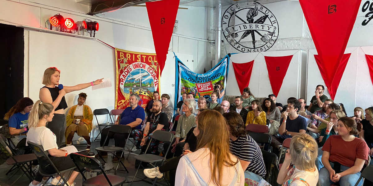 A speaker with a microphone addresses a packed room in an assembly. Union banners hang from the wall behind