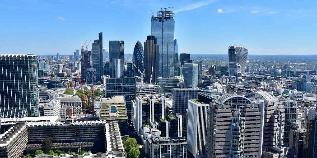 A City of London skyline photo taken from high up, with parts of the Barbican estate visible to the left and glass towers including the Gherkin and 'Walkie talkie' further in the distance against a bright blue sky