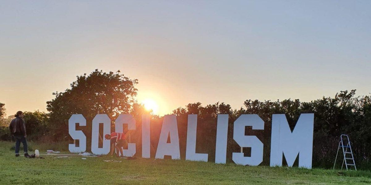 Large white letters spelling out 'SOCIALISM' stand against a hedgerow in a field at daybreak, with figures in front securing the letters