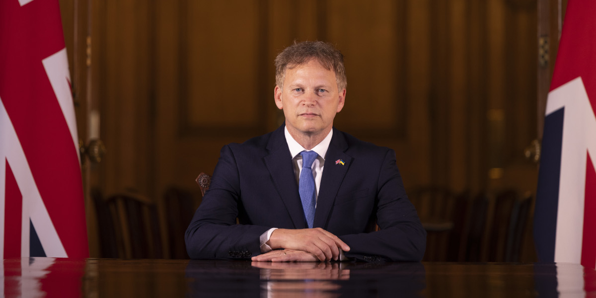 Official photo of Grant Shapps sitting at a table in front of British flags