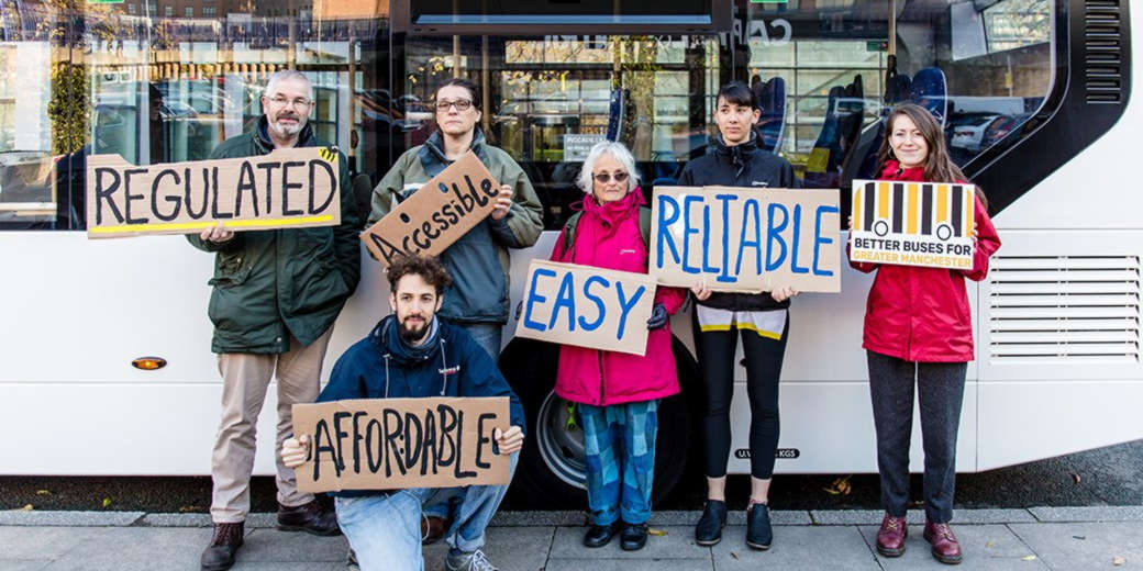 Six people stand in front of a bus holding signs with simple, handwritten slogans in support of public ownership of buses.