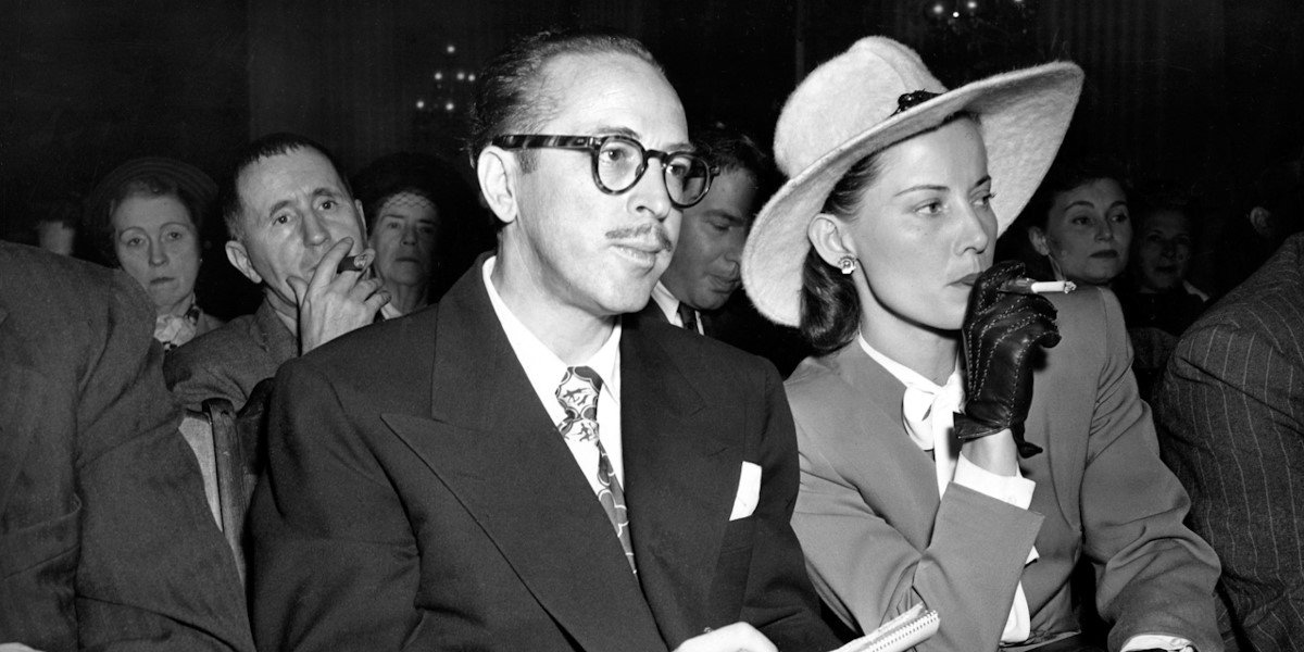 A black and white photograph. Screenwriter Dalton Trumbo sits next to his wife Cleo at the House Un-American Activities Committee hearings in 1947. Bertoldt Brecht sits in the background