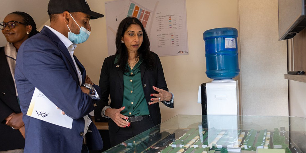 Suella Braverman speaks to a man wearing a face mask in Rwanda. They are looking at a small model of a building.