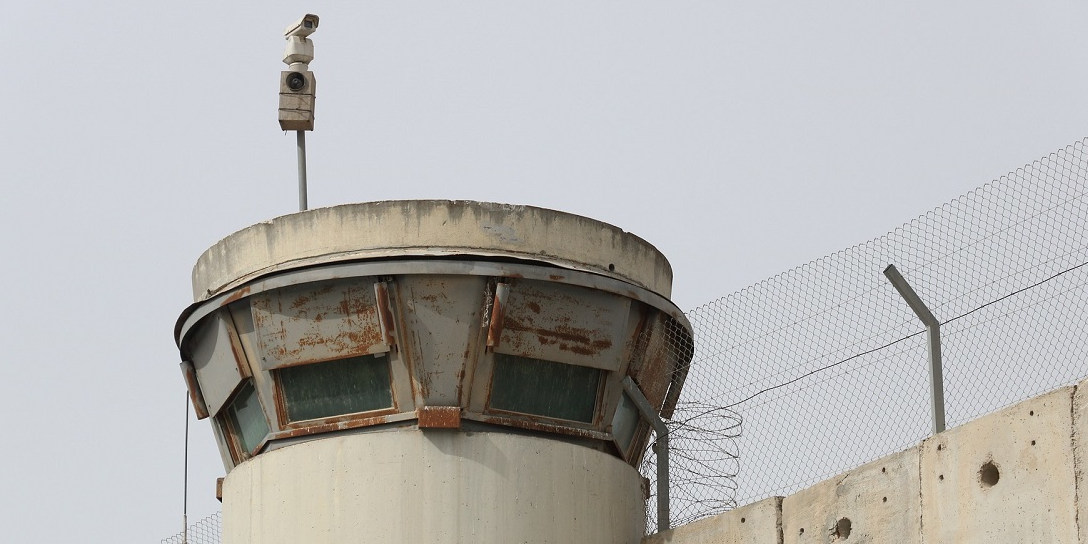 The image depicts an Israeli border crossing checkpoint with high barbed wire and a central look out tower