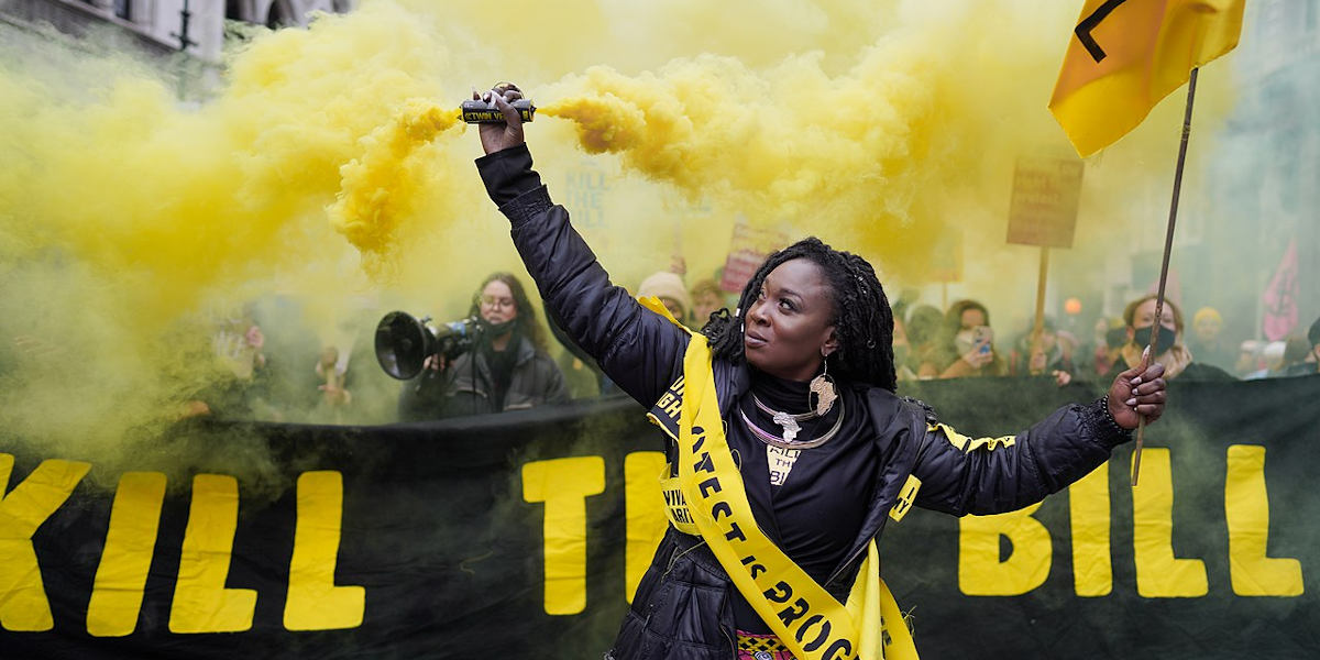 A woman at a protest waiving a flare billowing yellow smoke in front of a banner reading "Kill the Bill"