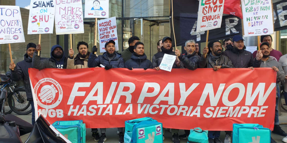 Deliveroo riders stand behind a banner that reads 'Fair Pay Now' with the IWGB logo. Their deliveroo food bags are in front of them. They are holding placards.