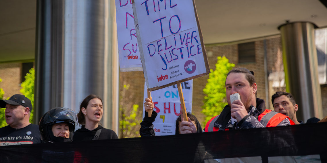 Deliveroo riders at a protest hold up placards. One driver speaks into a megaphone.