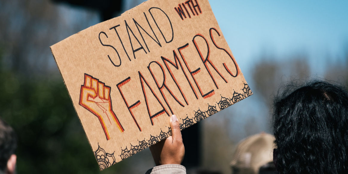 A hand holds a cardboard sign that says 'Stand with Farmers' and has a drawing of a raised fist. We see the back of somebodies head with black hair. 