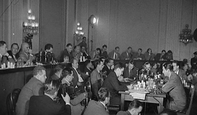 A black and white photograph of a committee room. There are lots of men in suits