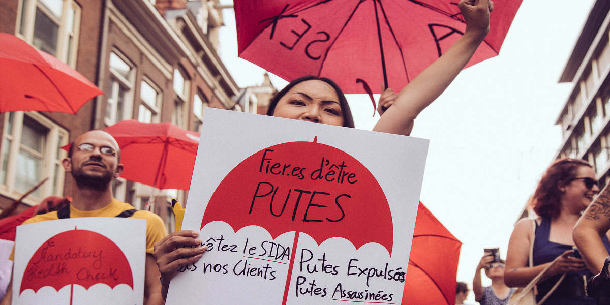 Protestors will placards and red umbrellas. The central placard reads 'proud to be whores' in French