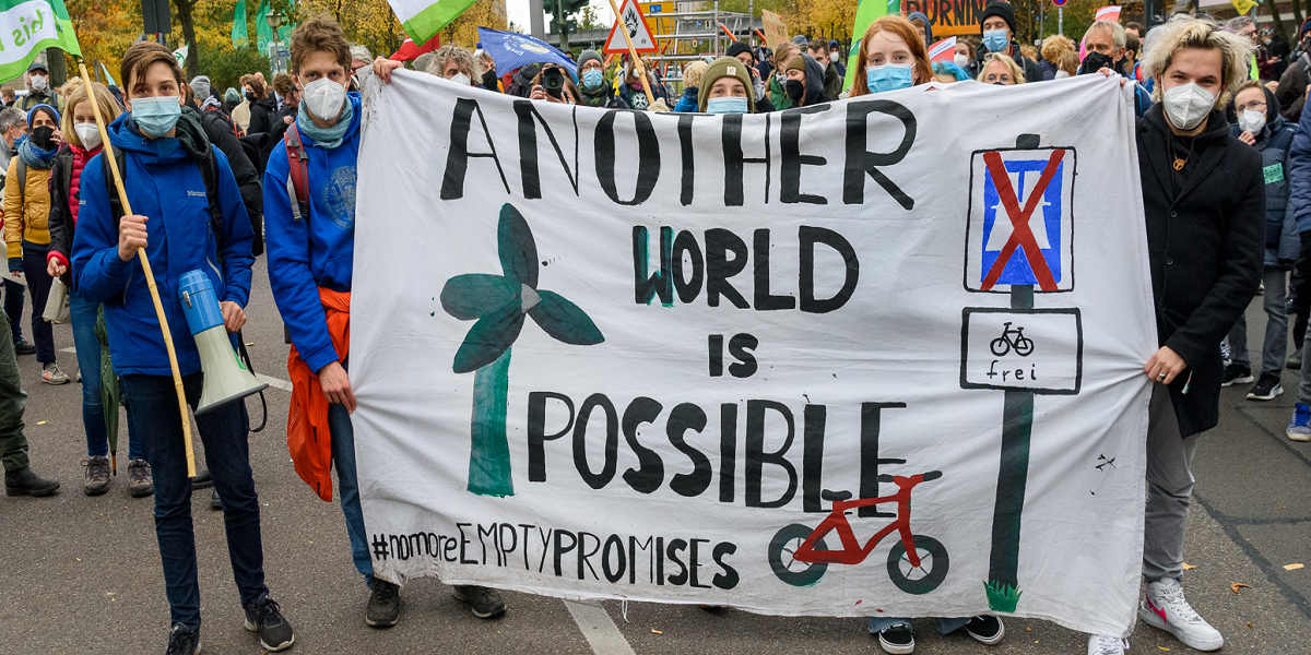 A crowd of young people hold up a large hand painted banner that reads 'Another world is possible' with a wind turbine and bike painted on it, with the hashtag slogan: 'No more empty promises' at the bottom