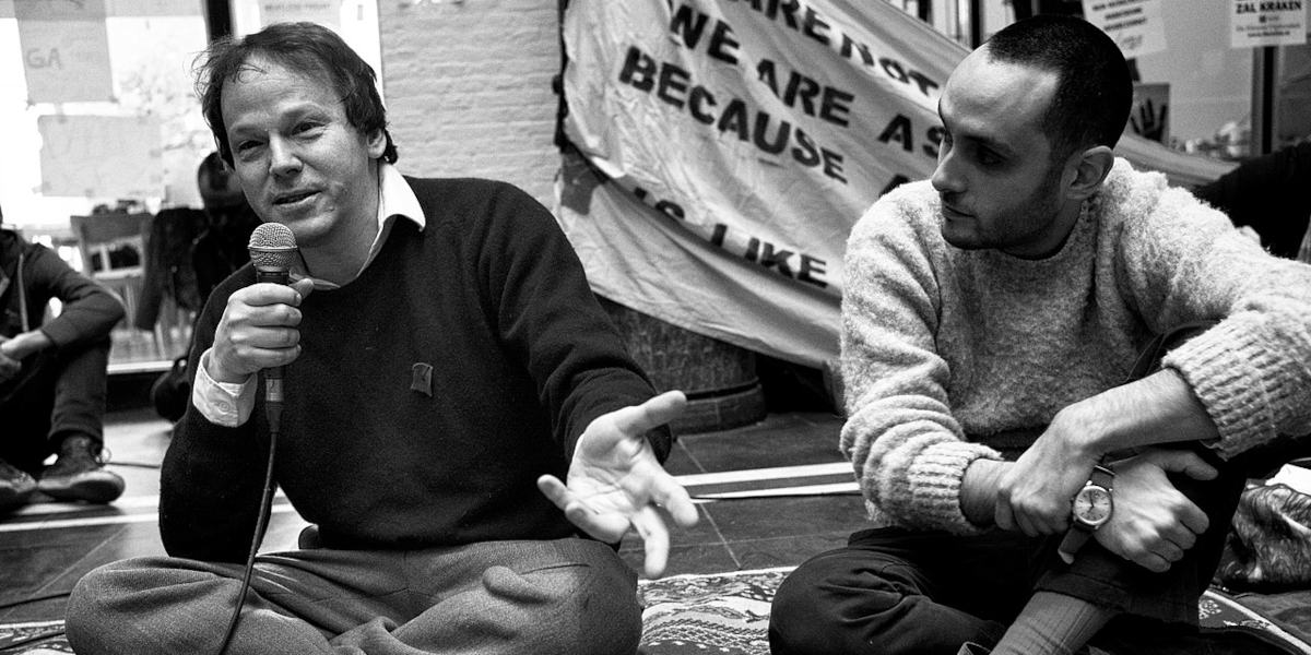 A black and white photo shows two men sitting crossed legged on the floor, a protest banner in the background. The man on the left is David Graeber, holding a microphone and gesturing to unseen listeners