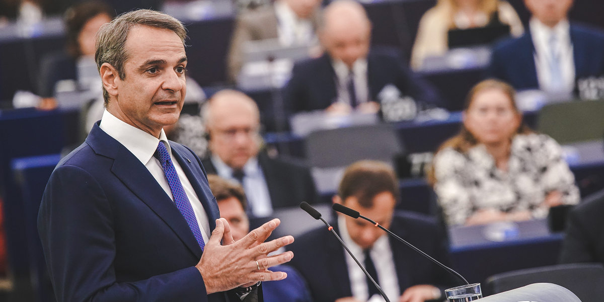 Kyriakos Mitsotakis, a white man wearing a suit with his hands clasped, speaks into a microphone against a backdrop of more white men in suits listening from their seats in the European Parliament