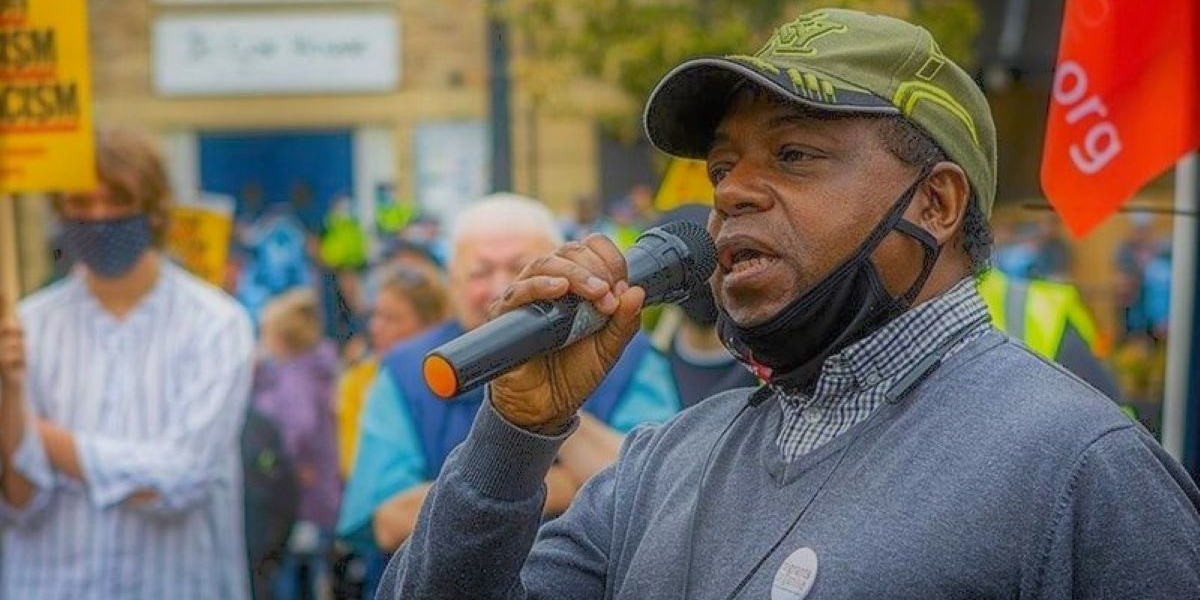 Against a backdrop of a rally crowd, Sarli Nana holds a microphone up to his face to make a speech, wearing a cap and with a face mask pulled down under his chin