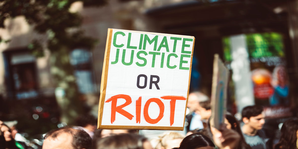 A placard reading 'Climate Justice or Riot' is held up above a sea of heads at a protest rally