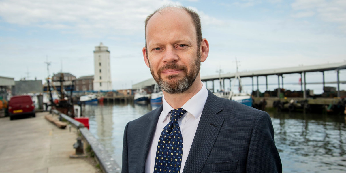 A portrait of a white man in a suit standing in front of a river in an industrial setting