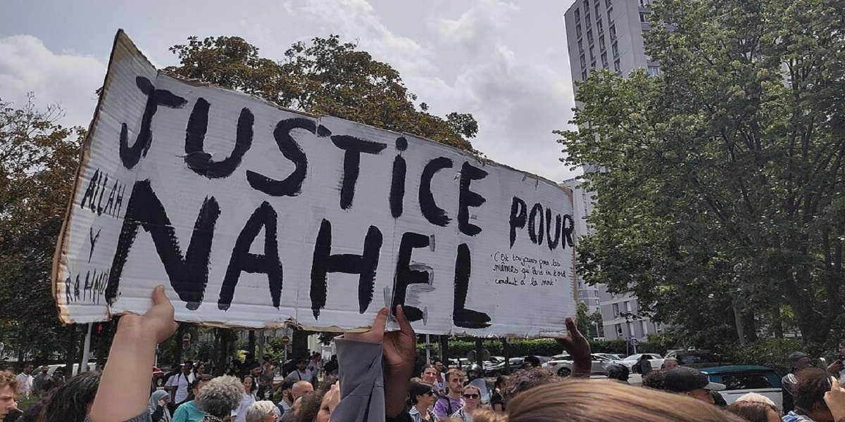 A hand-painted placard that reads 'Justice our Nahel' is held up in the air by protestors at a rally