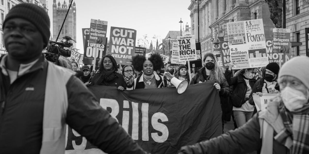 Protestors marching with banners reading 'cut war not welfare' and other references to the cost-of-living crisis. In the foreground, a black man and an older white woman in a mask link arms