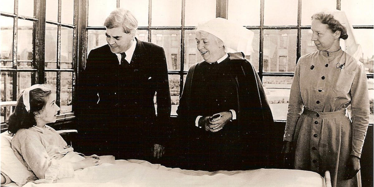A child lies in a hospital bed, alert and smiling up at a man, Aneurin Bevan, and two women dressed in nurses uniforms typical of the 1940s, on the first day of the National Health Service