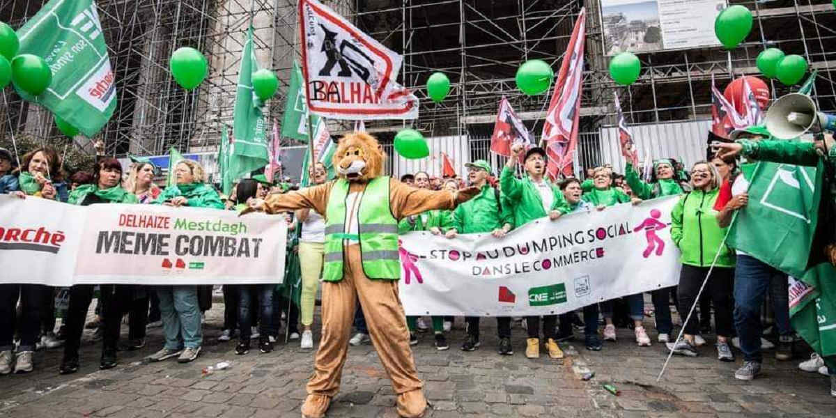 A CNE trade union protest. Group of people in green jackets holding banners and green balloons. A person in a lion suit in a hi-vis jacket stands in front with arms outstretched.