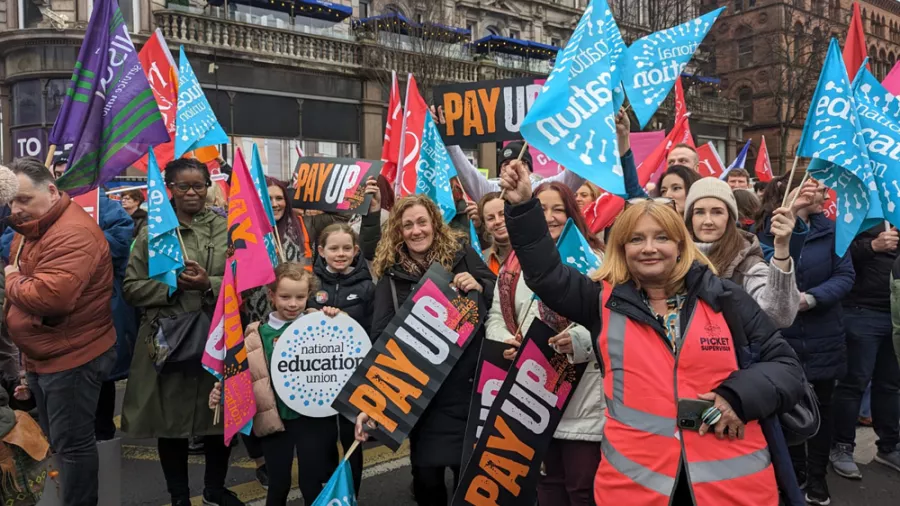 Teachers and pupils hold up National Education Union flags on the picket line. They are holding signs that say Pay Up.