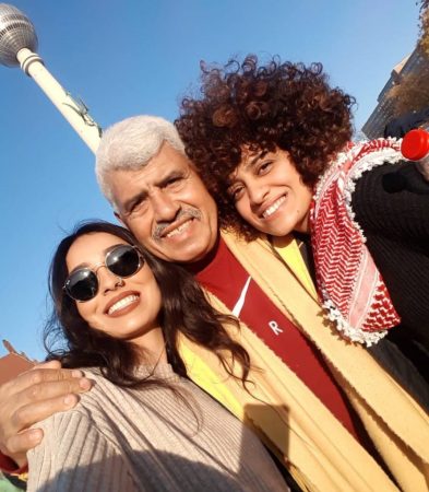 Ismail Abusalama stands between his two daughters hugging them, all are smiling against a blue sky city background